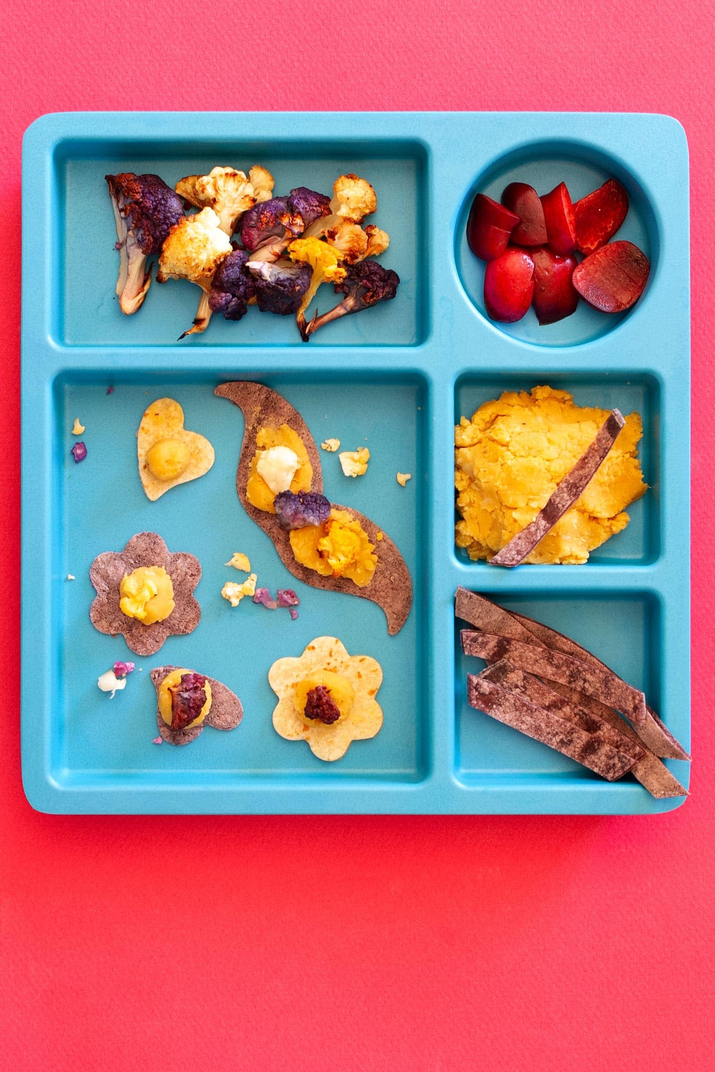 Top-down view of a blue bamboo kids plate filled with roasted purple and orange cauliflower, slices of fresh cherries, cooked red lentils, purple tortilla strips, and mini tacos in the shape of hearts, flowers, and a mustache.
