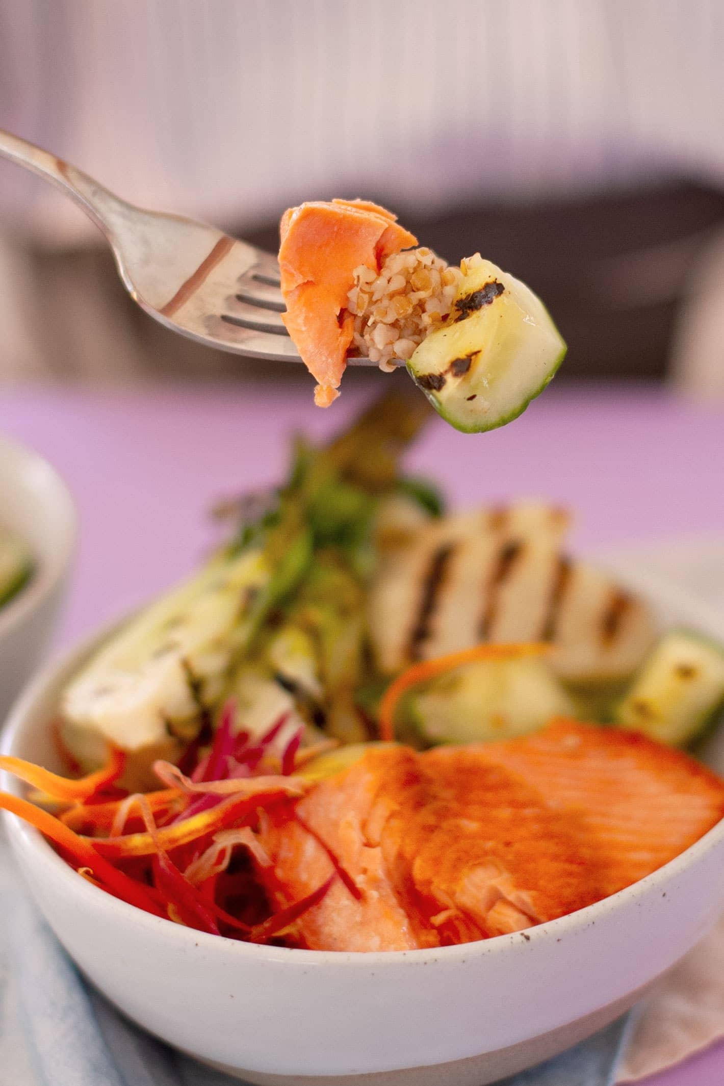Close-up view of a piece of salmon, bulgur, and grilled cucumber on a fork lifted above a bowl of pan-seared salmon, carrot strips, and grilled vegetables.
