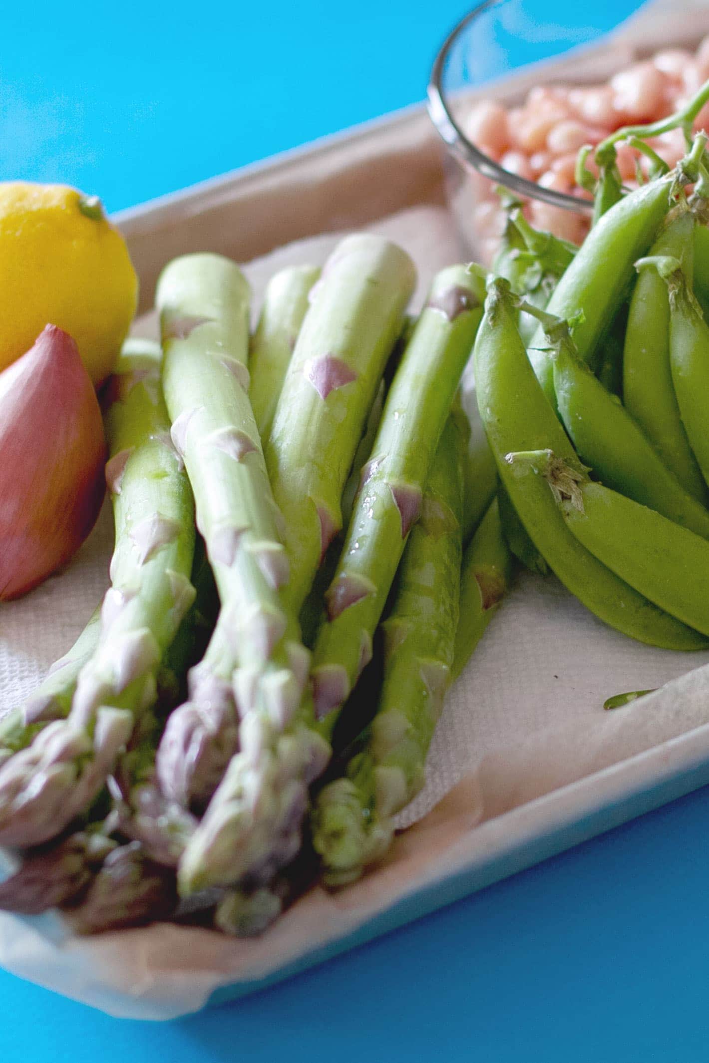 Fresh asparagus stalks, sugar snap peas, a shallot and lemon, and a clear bowl of white beans displayed on a parchment paper covered small sheet tray over a bright blue surface.