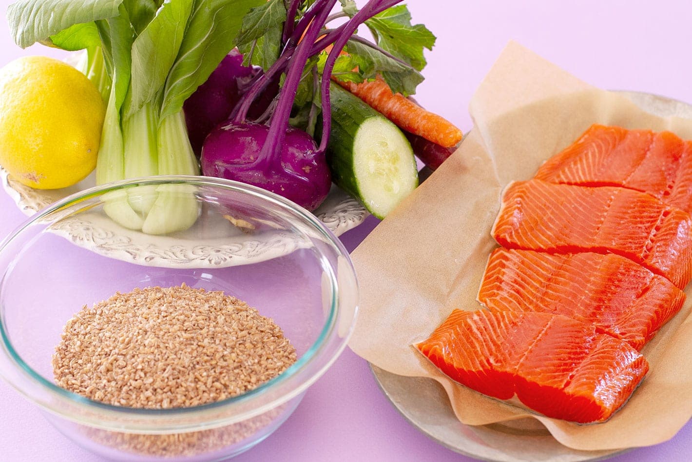 Ingredients for salmon grain bowl recipe sitting on a pastel purple surface - a bowl of bulgur, raw salmon fillets on a sheet tray, and fresh baby bok choy, kohlrabi, cucumber, carrots, and a lemon on a white plate.
