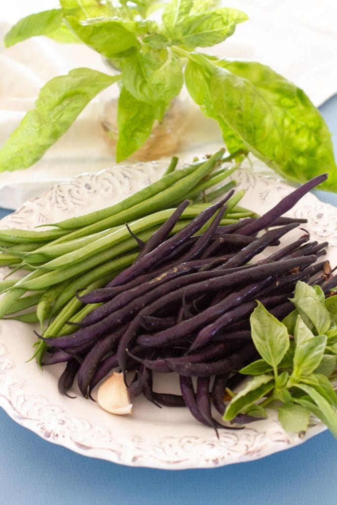 Vertical shot of fresh cleaned green and purple string beans, a glove of garlic and fresh basil on a white plate over soft blue paper. A basil plant in vase is in the background.