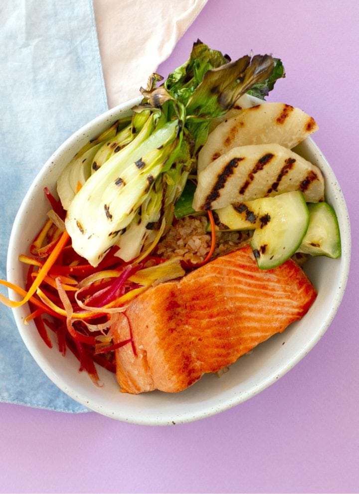 salmon bulgur grain bowl with grilled vegetables in a white ceramic bowl on a light blue washed fabric and violet paper