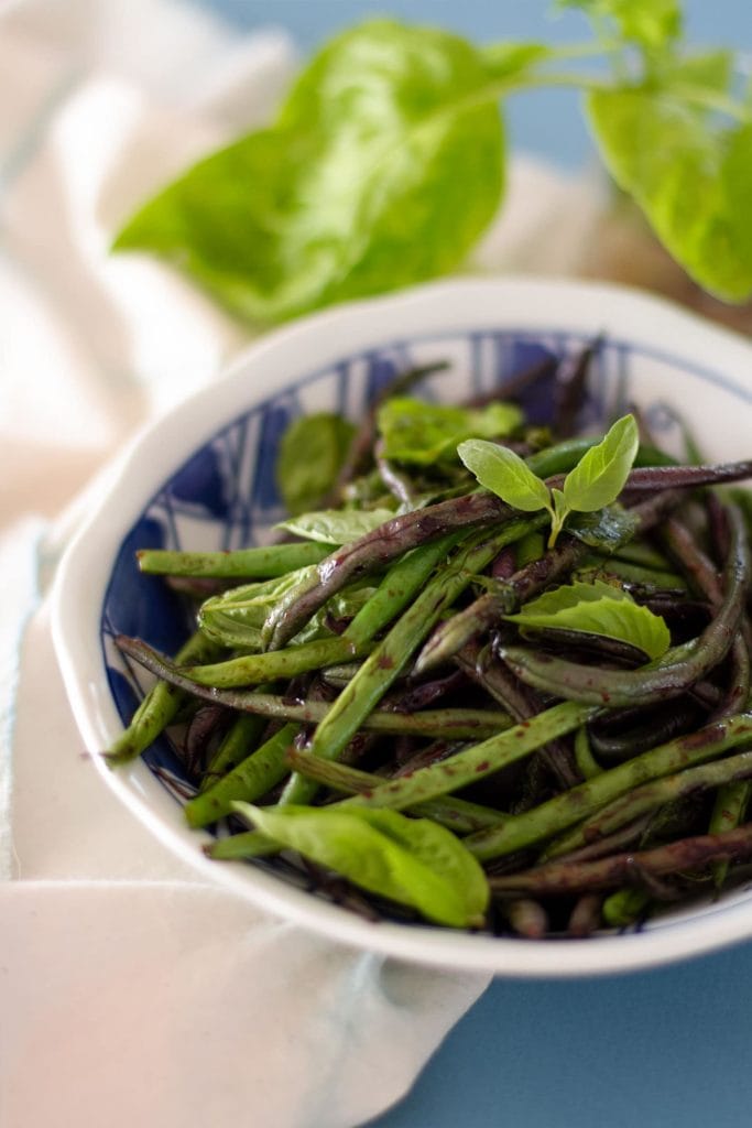 ¾ angle shot of sauteed green beans topped with fresh basil in a white plate over a white cloth and soft blue paper.