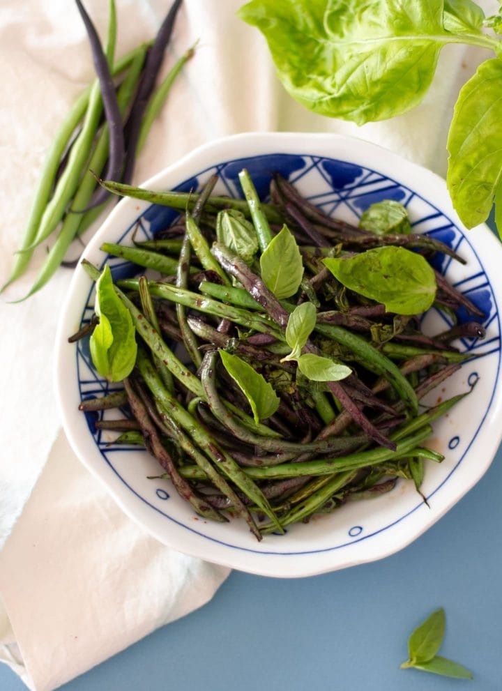 Top down view of sauteed green beans with fresh basil in a white patterned bowl over a soft blue background and fresh green and purple beans on the side.