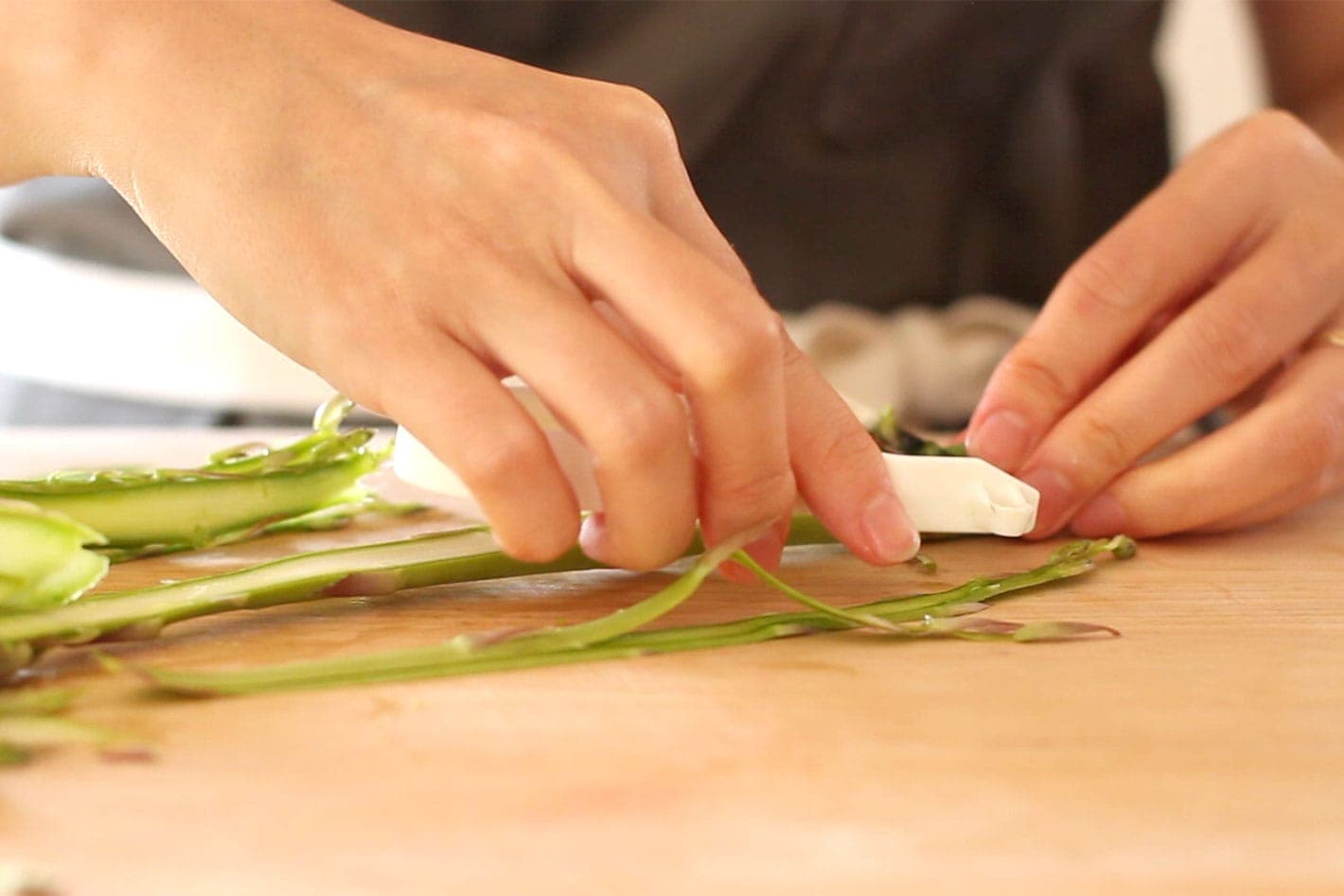 Raw asparagus on a wood cutting board being shaved into thin strips with a white vegetable peeler.