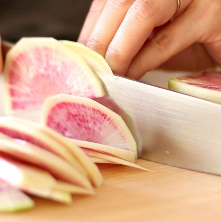 Closeup view of watermelon radish being sliced into thin, half-mooned pieces.