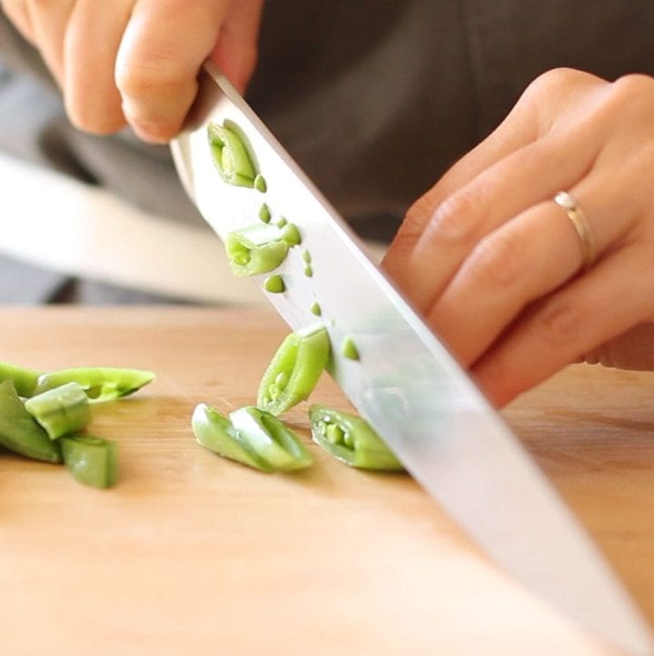Close-up angle view of sugar snap peas being sliced on the bias over a wood cutting board.
