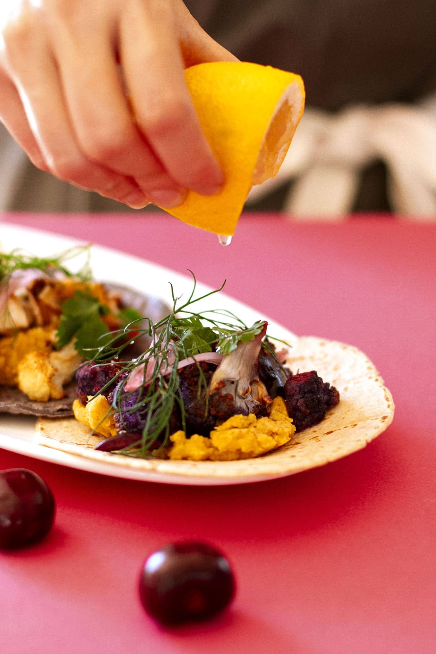 Close up view of a hand squeezing lemon juice over roasted cauliflower and lentils tacos topped with fresh cherries and dill. 