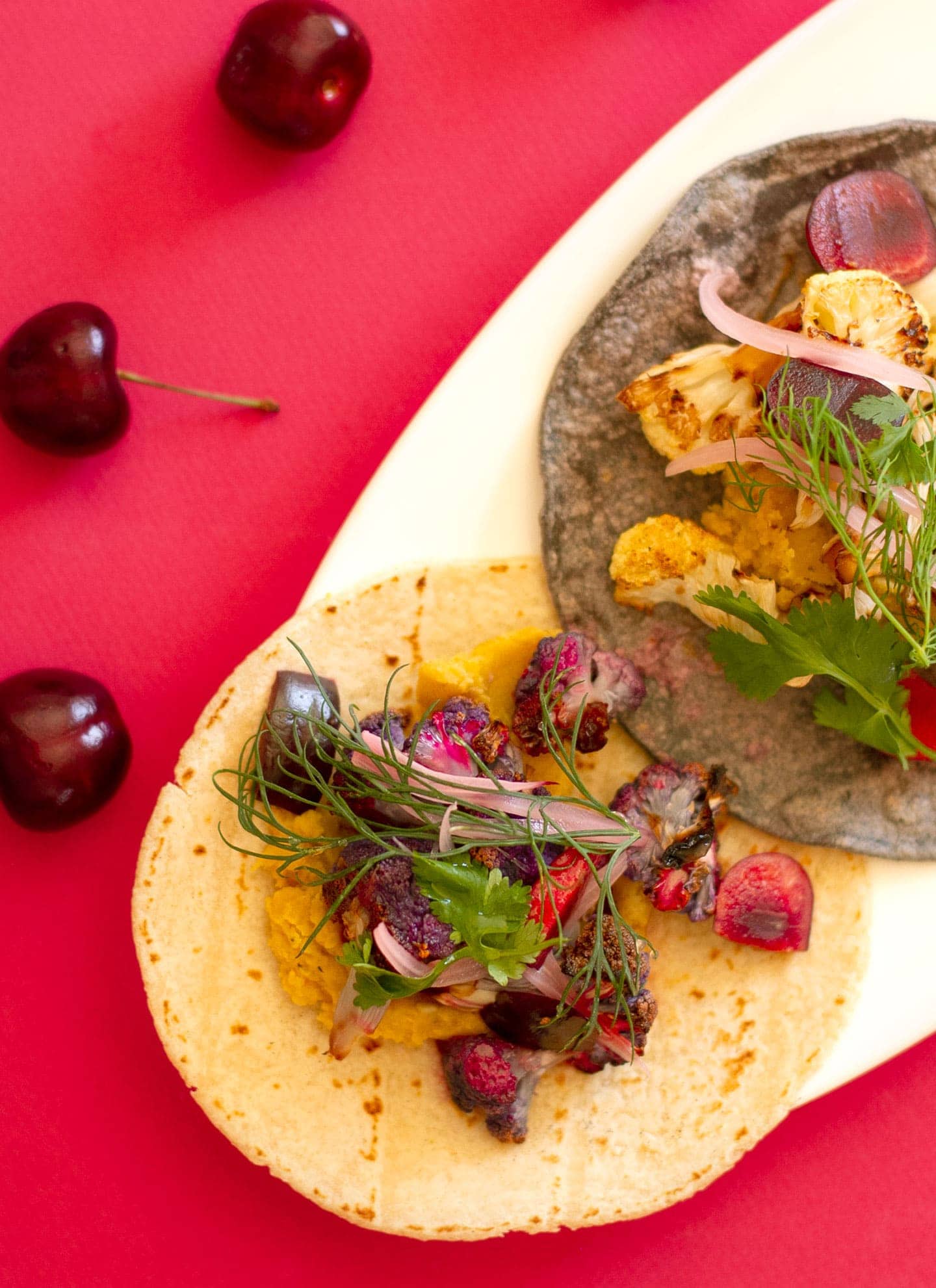 Top down view of tacos filled with roasted cauliflower, lentils, and fresh cherries on a white plate over a bright red background surface.