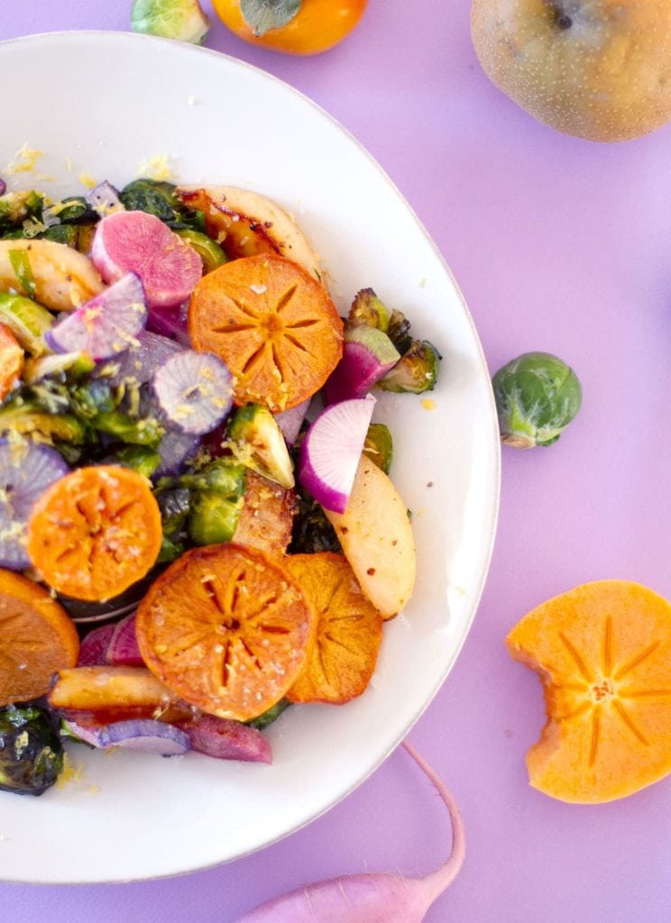 A plate of colorful roasted persimmon, brussels sprouts, pear, and pear with the raw ingredients surrounding the finished dish.