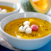 korean pumpkin porridge topped with sweet rice balls and black sesame seeds in a white bowl with a slice of raw kabocha in the background