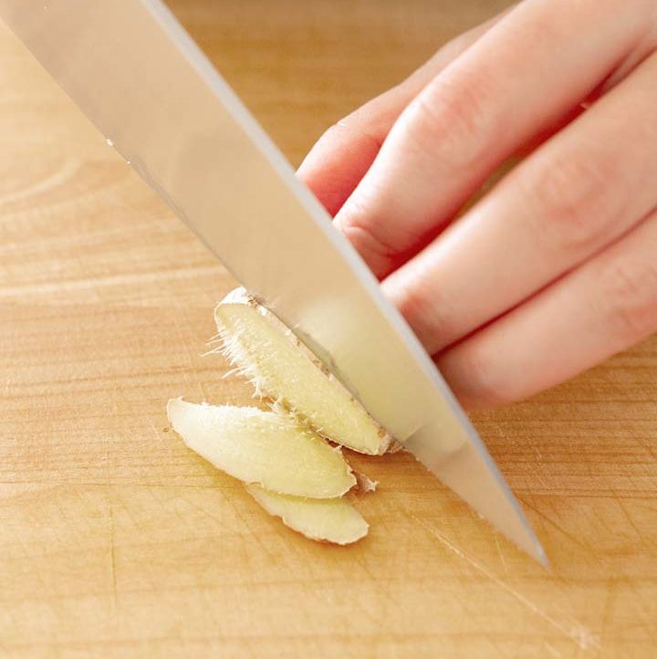 top view of ginger being thinly sliced with a chef knife on a wood cutting board