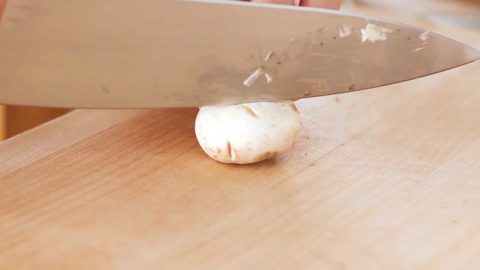 White button mushroom on a wood cutting board with a chefs knife ready to slice.