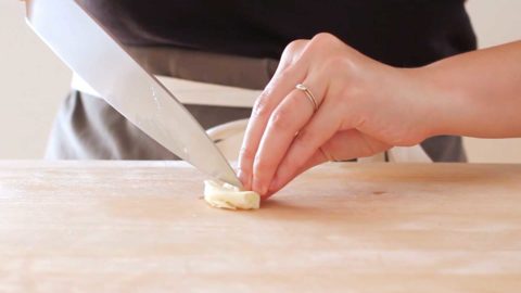 A hand holding a smashed clove of garlic on a wood cutting board while slicing with a chefs knife.