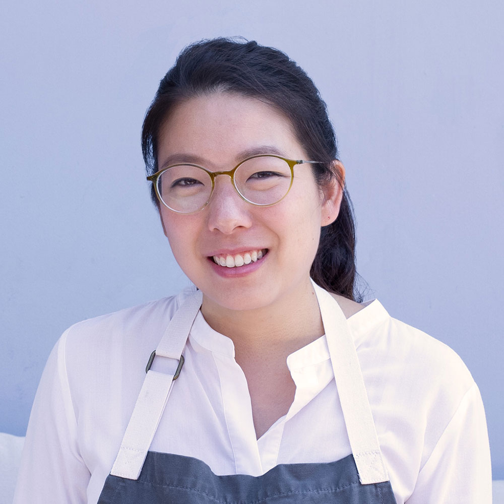 Headshot of Cindy smiling while wearing a white shirt and gray chef's apron standing in front of a blue background.
