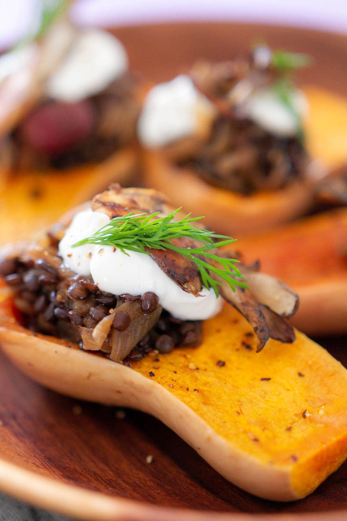 A close-up photo of roasted honeynut squash filled with black lentil stuffing, yogurt, dill, and seared maitake mushroom on a wood plate.
