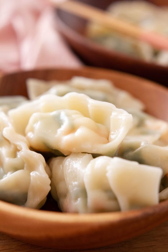 A bowl of boiled dumplings filled with snow pea leaves and shrimp with a pink cloth and another bowl with chopsticks in the background.