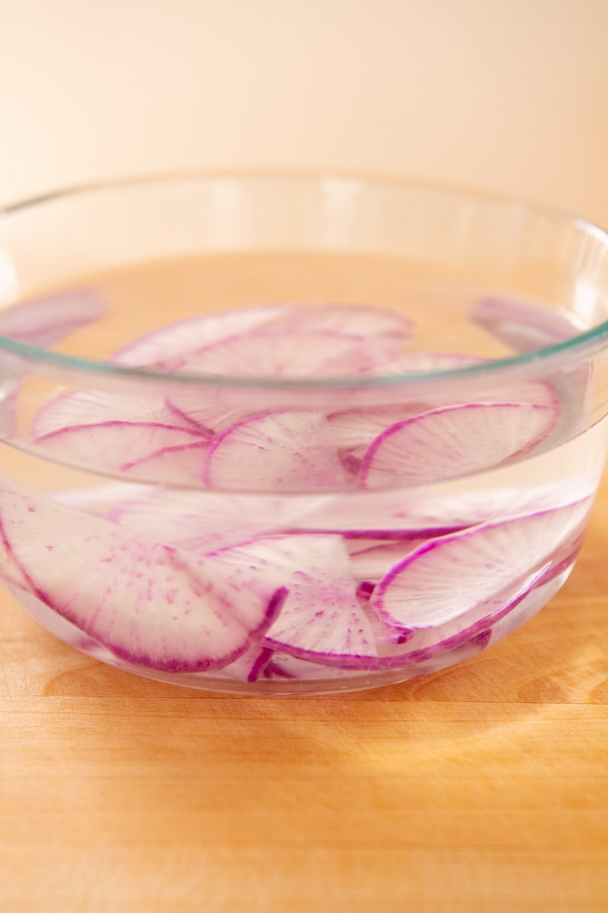 Thinly sliced purple daikon submerged in water in a clear bowl that's sitting on a wood cutting board.