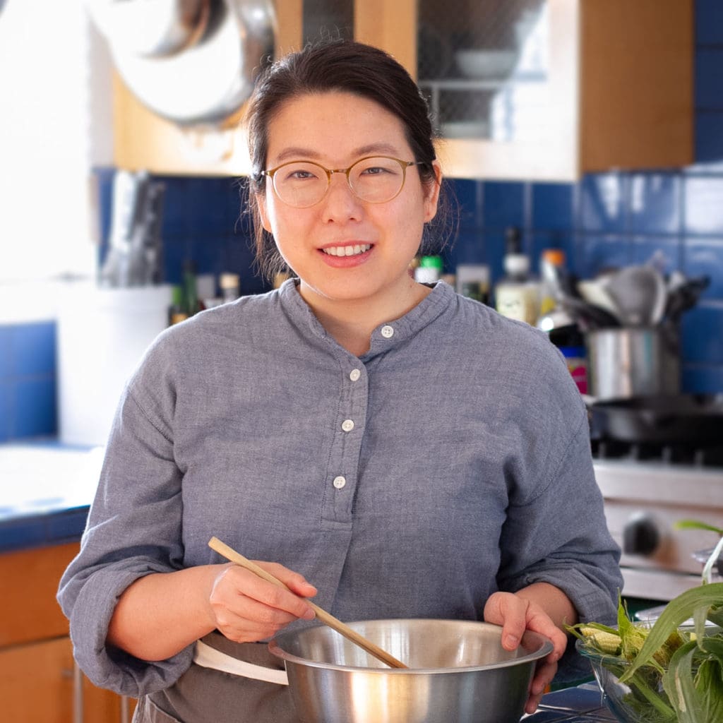 Cindy smiling while wearing a blue chefs coat and gray apron, holding chopsticks and a mixing bowl.