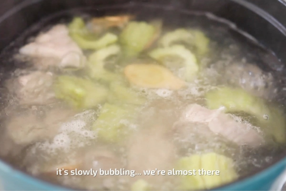 A pot of simmering bitter melon soup with pork ribs and text that reads, "it's slowly bubbling... we're almost there."