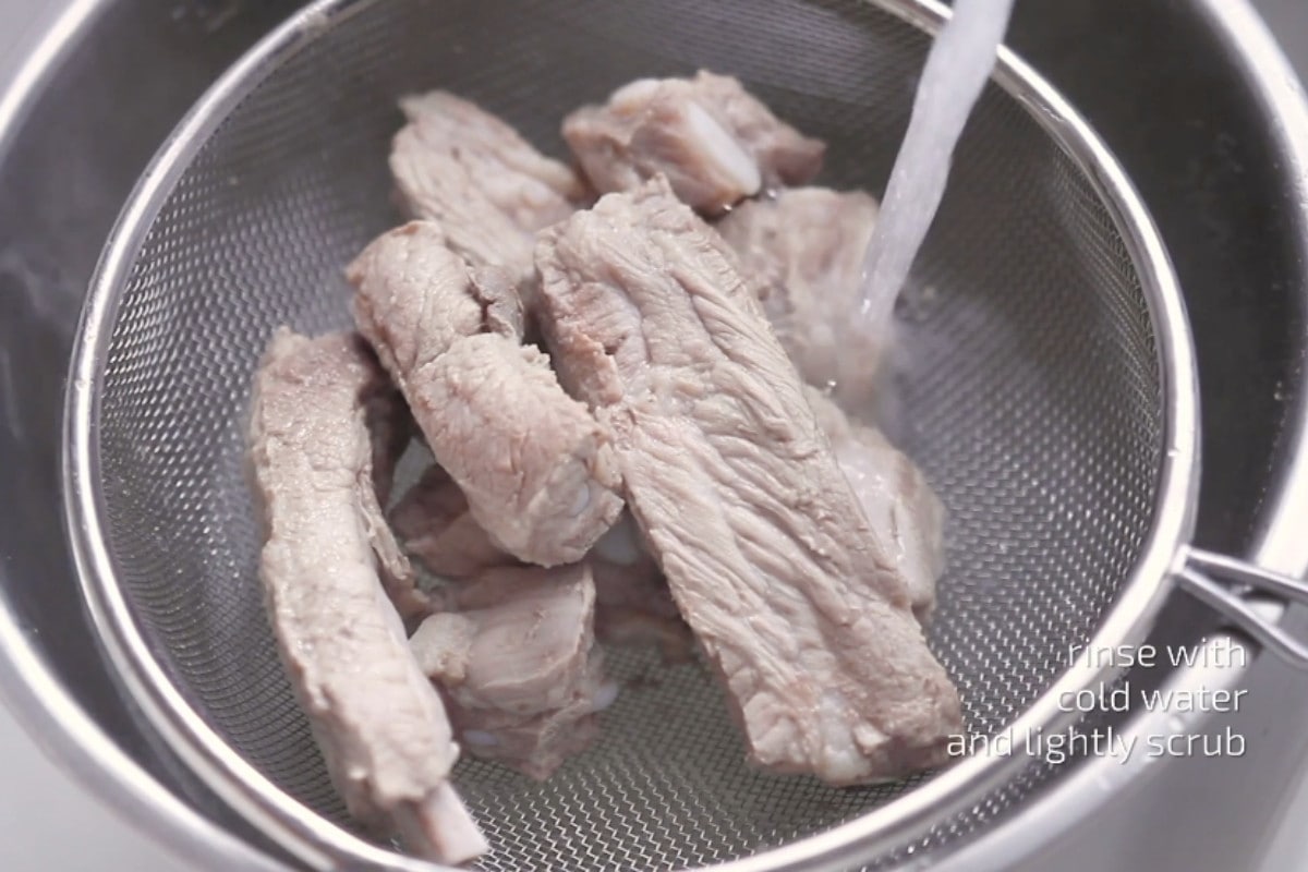 Blanched pork ribs in a strainer with water running over the ribs and text that reads, "rinse with cold water and lightly scrub."