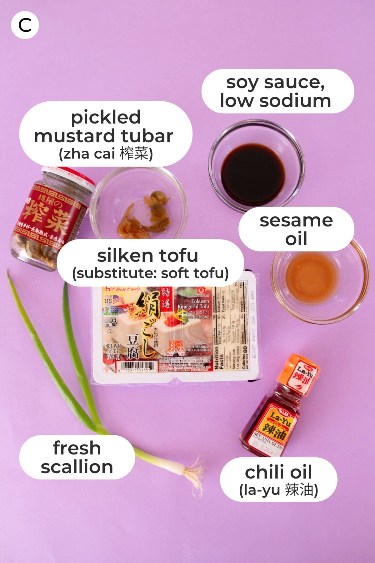 Ingredients of silken tofu with Japanese chili oil topping laid out over a purple background and labeled with a "C" as well as the ingredient names: pickled mustard tubar (zha cai 榨菜), sesame oil, silken tofu (substitute: soft tofu), sesame oil, fresh scallion, and chili oil (la-yu 辣油).