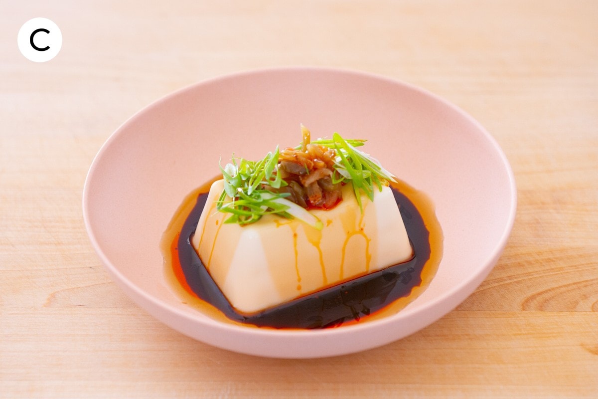 A photo labeled with "C" of silken tofu in a pink shallow bowl, topped with soy sauce, sesame oil, zha cai (pickled mustard tubar), and thinly sliced scallions.
