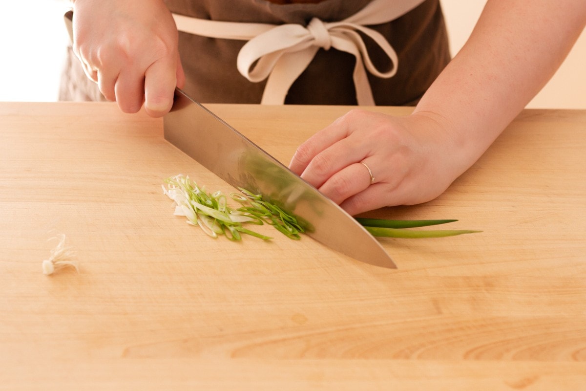Cindy thinly slicing scallion on a bias with a chefs knife on a wood cutting board.
