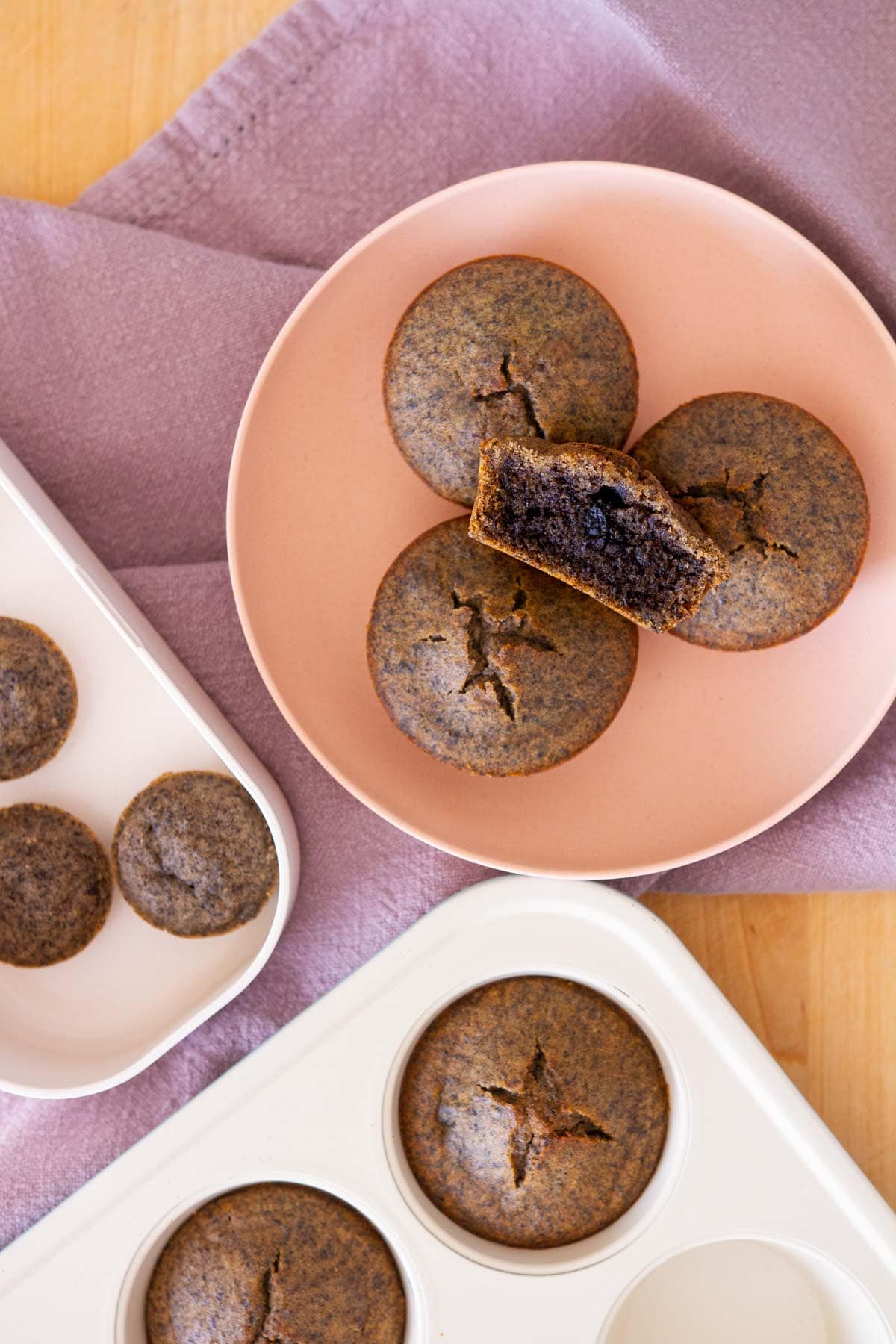 Top down view of stacked black sesame mochi muffins in a pink serving plate, with one cut in half to show the golden crust on the outside and moist center and mini ones on the side.