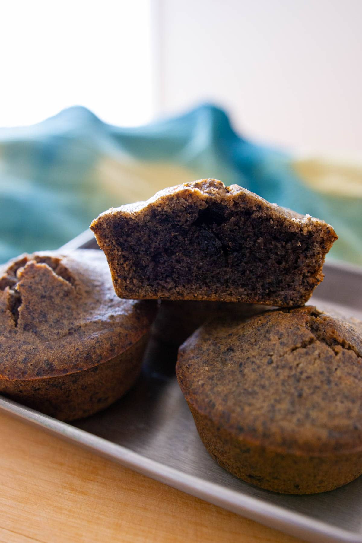 Close up view of stacked black sesame mochi muffins in a small baking tray, with the top one cut in half to show the golden crust on the outside and moist, black sesame-filled center.