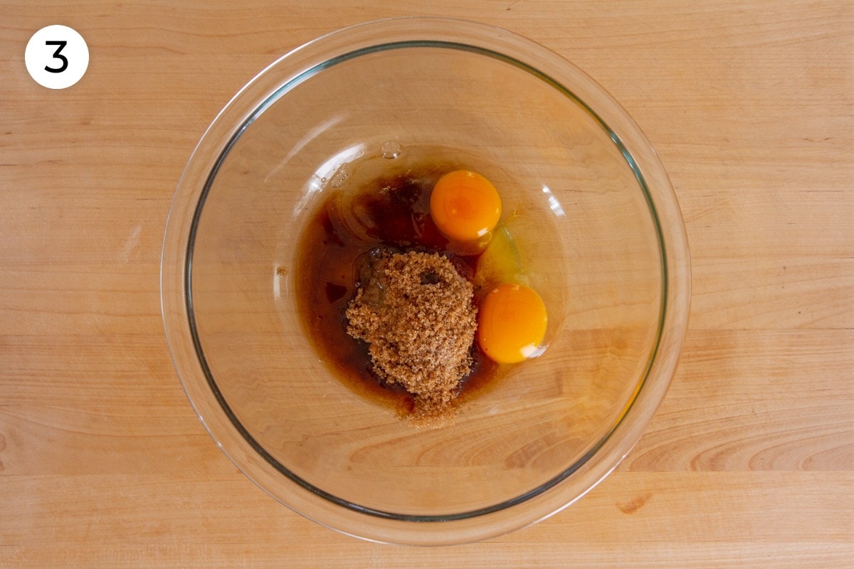 Light brown sugar, sugar, vanilla extract, and two eggs in a large glass mixing bowl on a wood surface, labeled with a circled number "3."