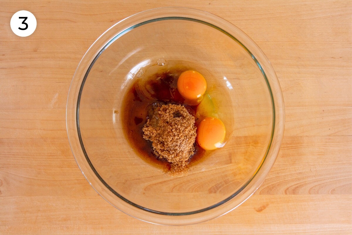 Light brown sugar, sugar, vanilla extract, and two eggs in a large glass mixing bowl on a wood surface, labeled with a circled number "3."