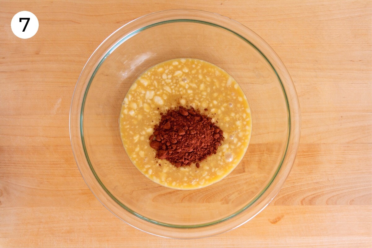Cocoa powder on top of the wet batter in a large glass mixing bowl on a wood surface, labeled with a circled number "7."