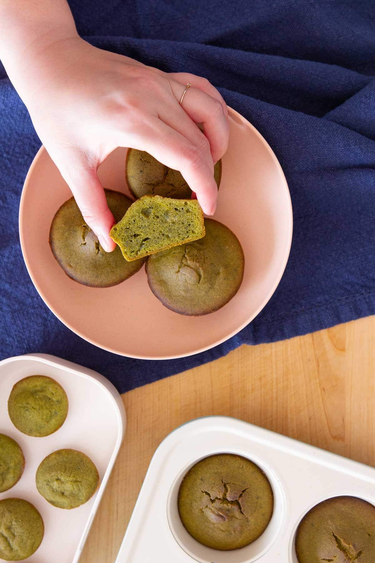 Top down view of stacked matcha mochi muffins in a pink serving plate, with a hand holding one cut in half to show the golden crust on the outside and moist center, along with muffins still in the pan and mini ones on the side.