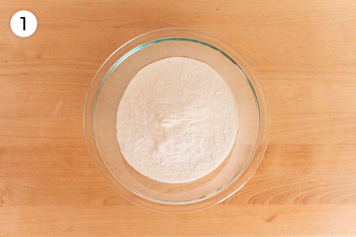 Dry ingredients (mochiko sweet rice flour, baking powder, and salt) in a medium glass mixing bowl on a wood surface, labeled with a circled number "1."