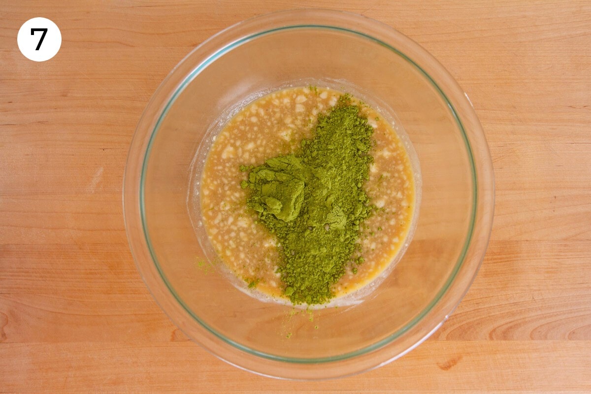 Matcha powder on top of the mochi moffin batter in a large glass mixing bowl on a wood surface, labeled with a circled number "7."
