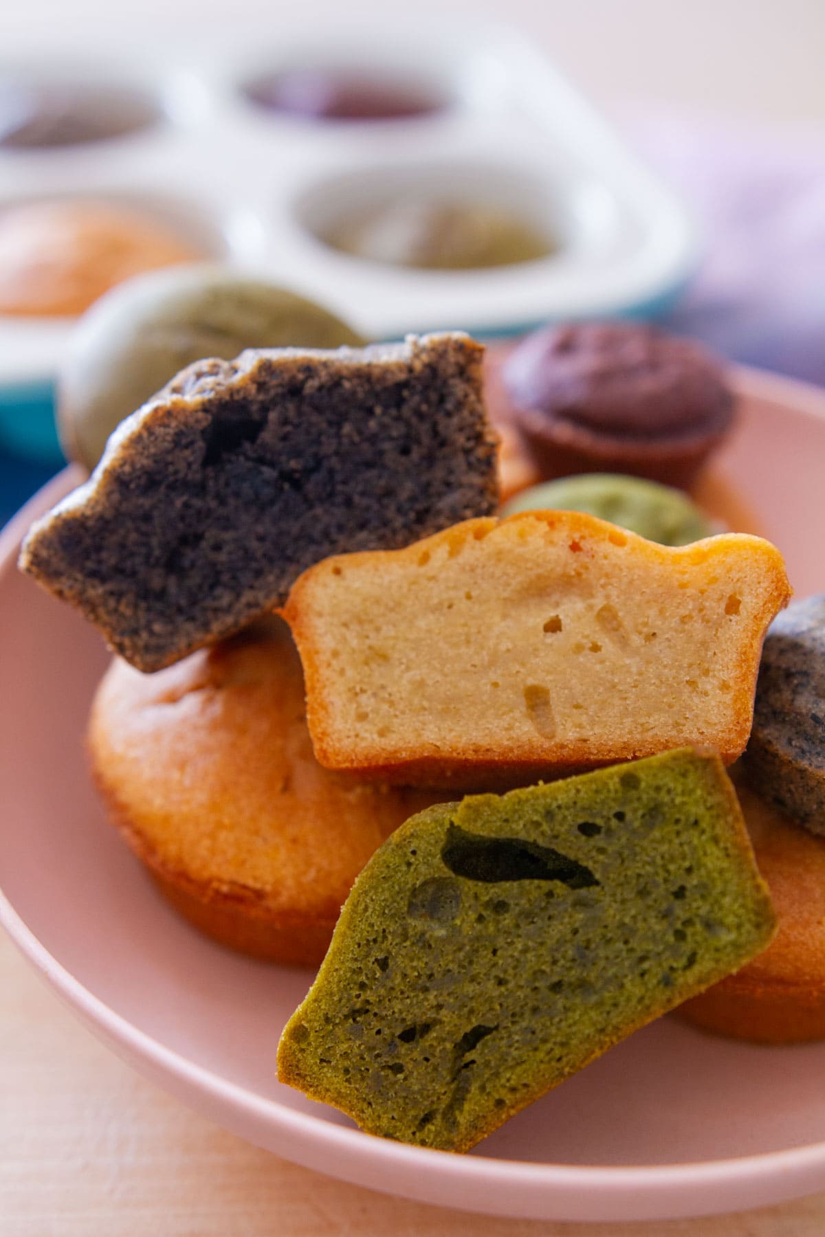 Close up view of three different mochi muffins cut in half to show the smooth texture on the inside, with the matcha flavor on the bottom, regular butter mochi flavor in the middle, and black sesame on top.