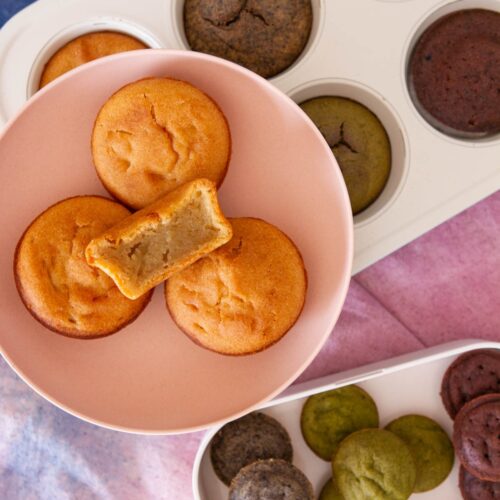 Mochi muffins in a small pink serving plate over different flavors (black sesame, matcha, and chocolate) of muffins still in the muffin pan and a container filled with a variety of mini mochi muffins.