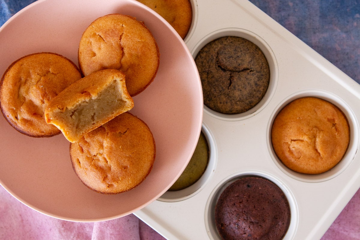 Mochi muffins in a small pink serving plate over different flavors (black sesame, matcha, and chocolate) of muffins still in the muffin pan and a container filled with a variety of mini mochi muffins.