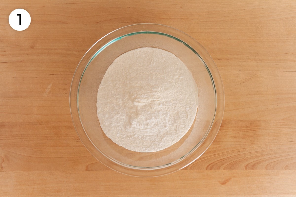 Dry ingredients (mochiko sweet rice flour, baking powder, and salt) in a medium glass mixing bowl on a wood surface, labeled with a circled number "1."