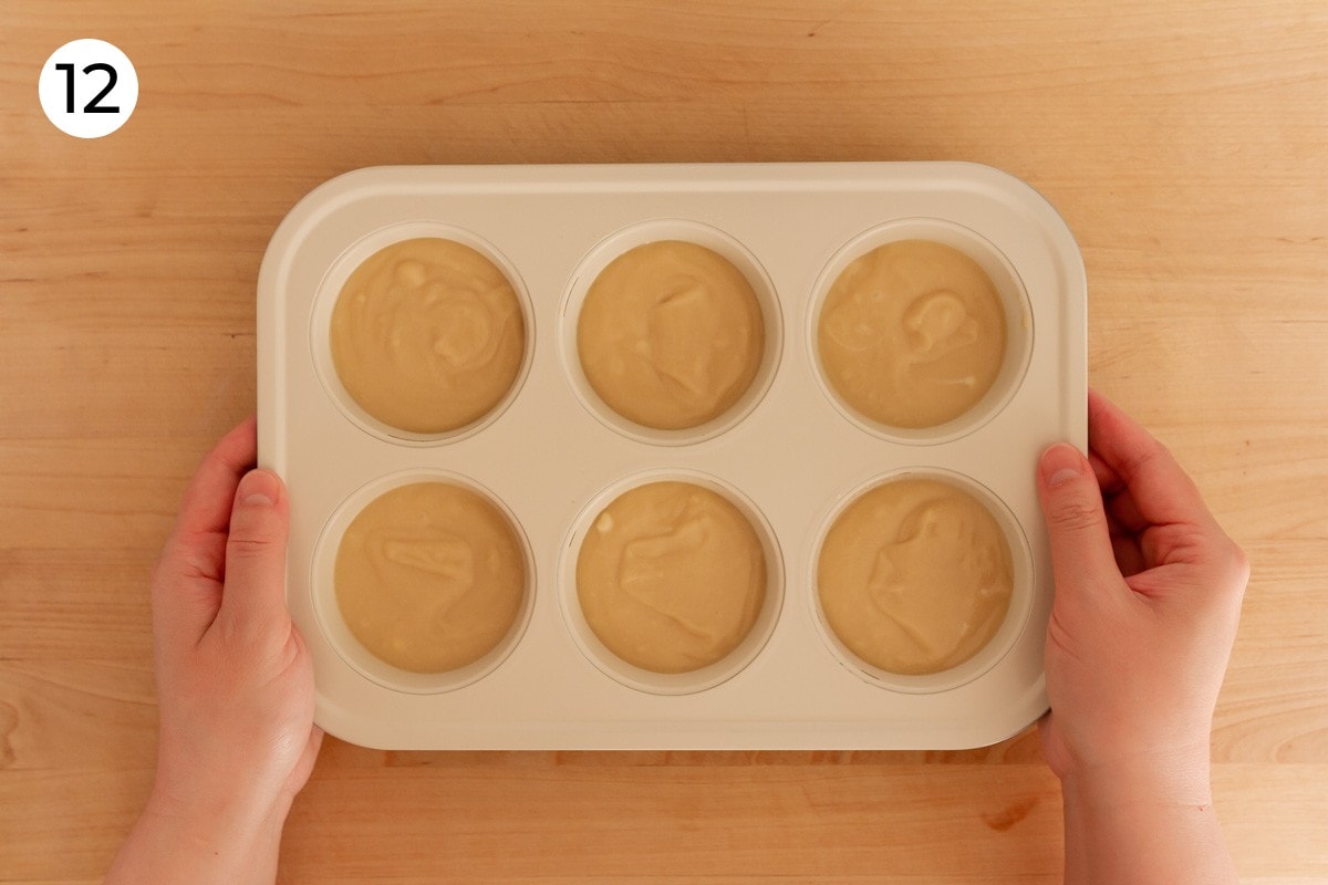 Cindy holding both sides of the filled muffin pan, tapping the bottom of the pan against a wood surface, labeled with a circled number "12."