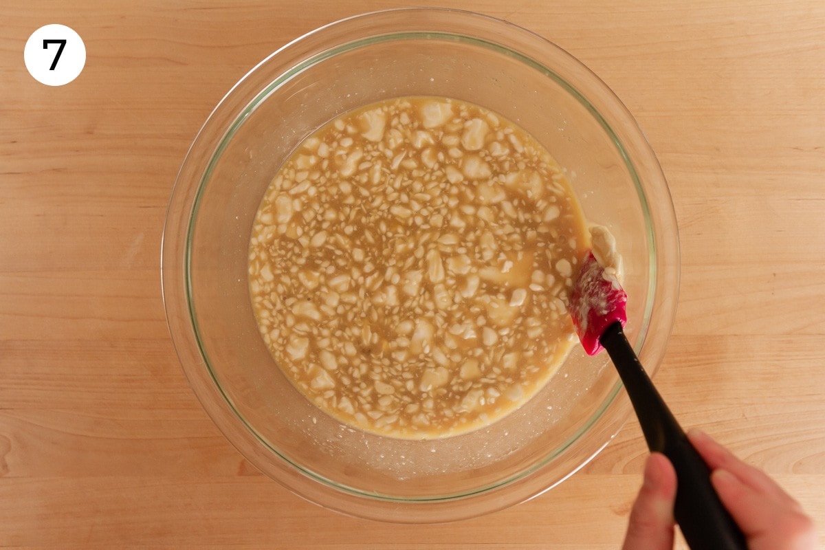 Cindy using a small rubber spatula to squeeze larger chunks of coconut milk fat against the sides of the large mixing bowl that's filled with the wet batter, labeled with a circled number "7."