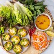 A bamboo serving tray filled with banh khot, assorted greens, dipping sauce, pickled carrots and daikon, slices of limes, and Thai chili peppers.