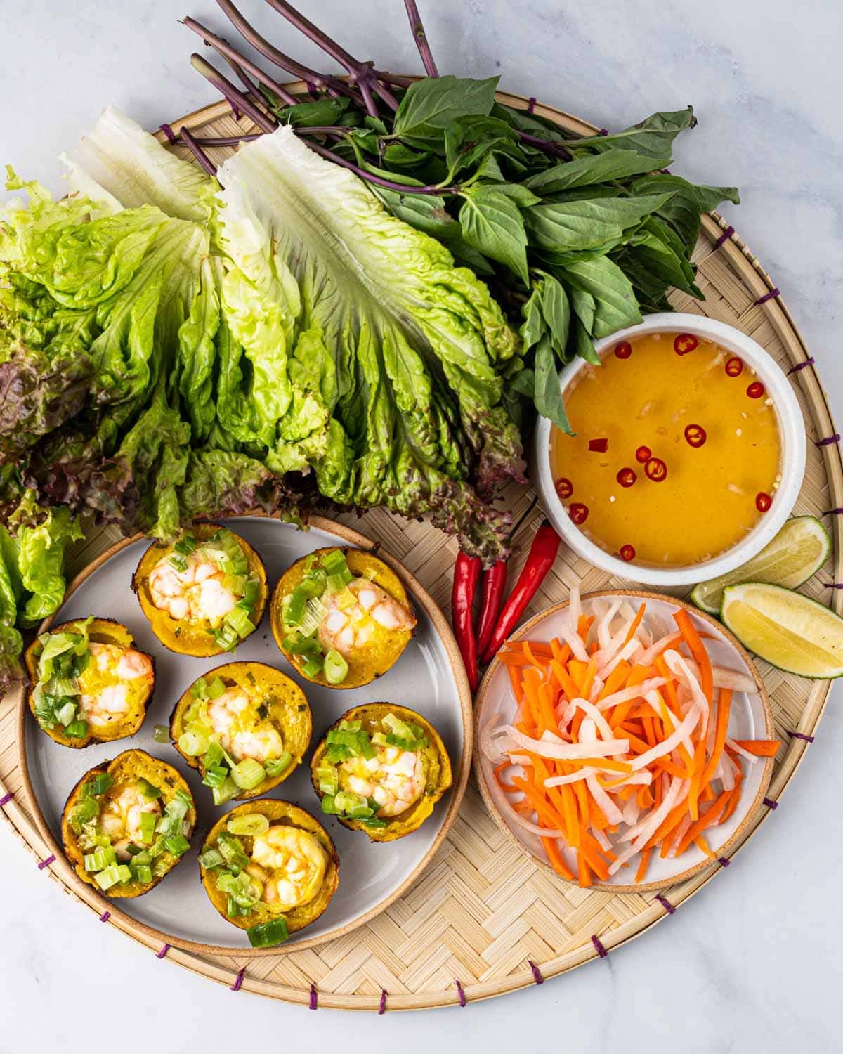 A bamboo serving tray filled with banh khot, assorted greens, dipping sauce, pickled carrots and daikon, slices of limes, and Thai chili peppers.