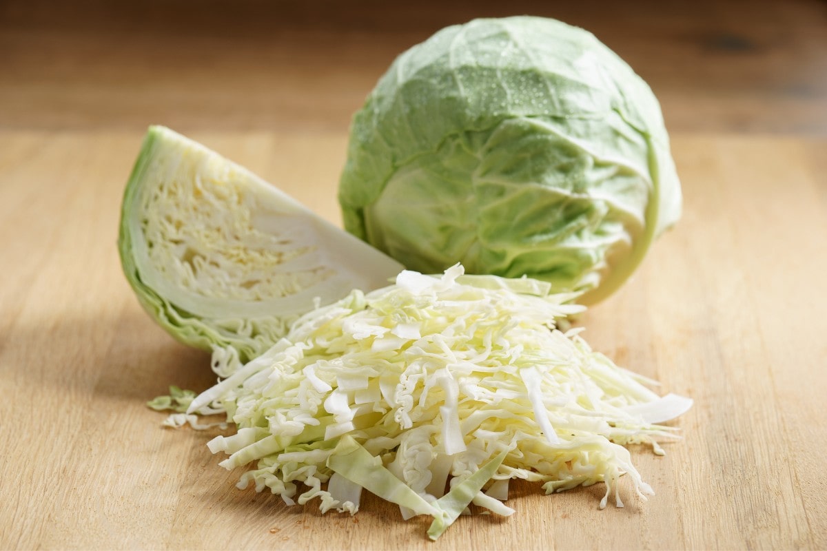 A pile of shredded cabbage in front of one head of green cabbage and a quarter of a cabbage on a wood cutting board.