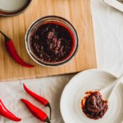 Open jar of homemade gochujang on a wood board surrounded by fresh red chilis.