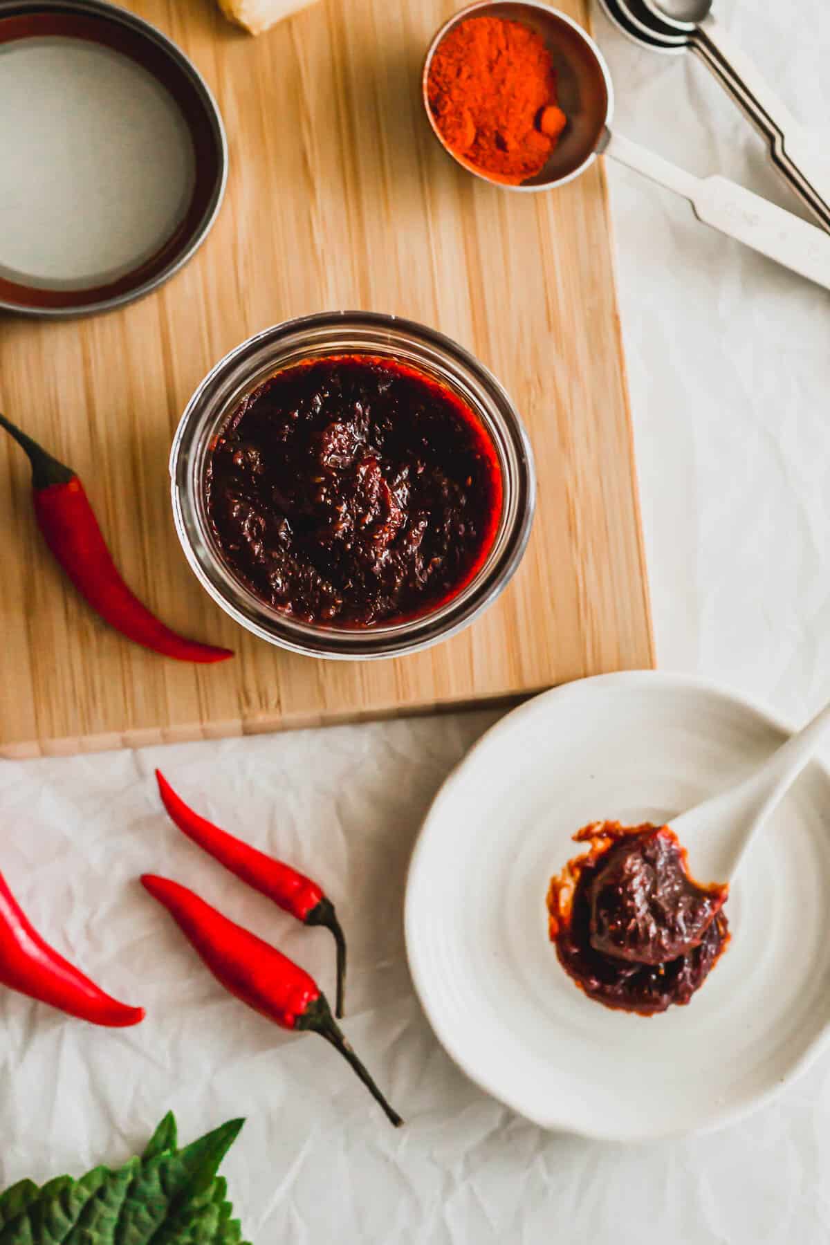 Open jar of homemade gochujang on a wood board surrounded by fresh red chilis.