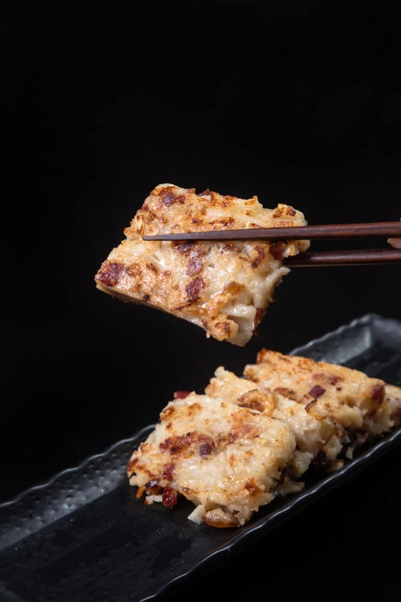 A pair of chopsticks holding up a piece of turnip cake over a plate lined with more.