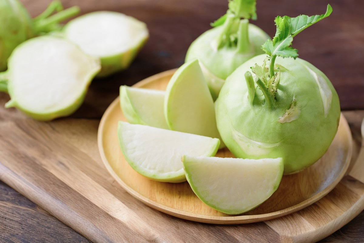 Two green kohlrabi next to slices of kohlrabi on a wood plate.