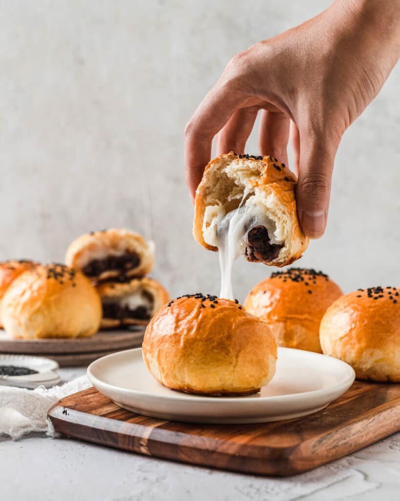 A mochi red bean bun cut in half on a white plate with a hand holding one half up to show the mochi pull.